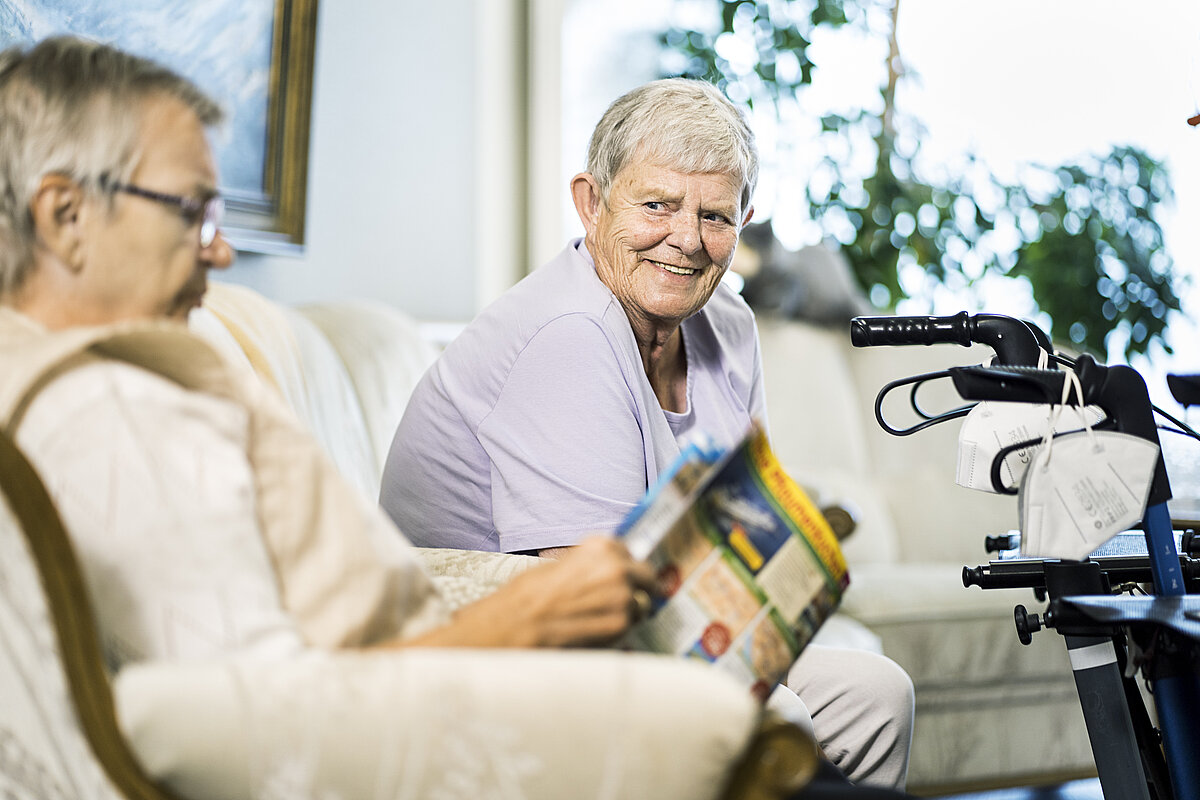 Bild zeigt zwei älteren Damen im Pflegeheim. Eine Damen  ließt eine Zeitschrift. Eine Dame schaut in die Kamera.